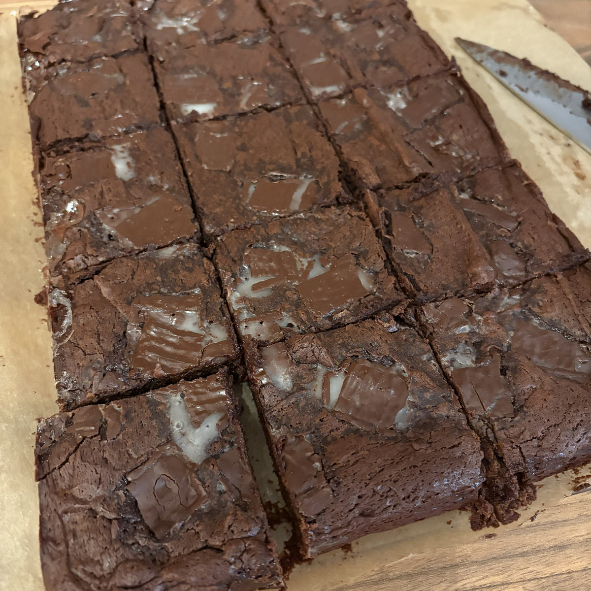A rectangular tray of freshly baked chocolate brownies cut into square portions, with some white icing on top, placed on parchment paper on a wooden surface.