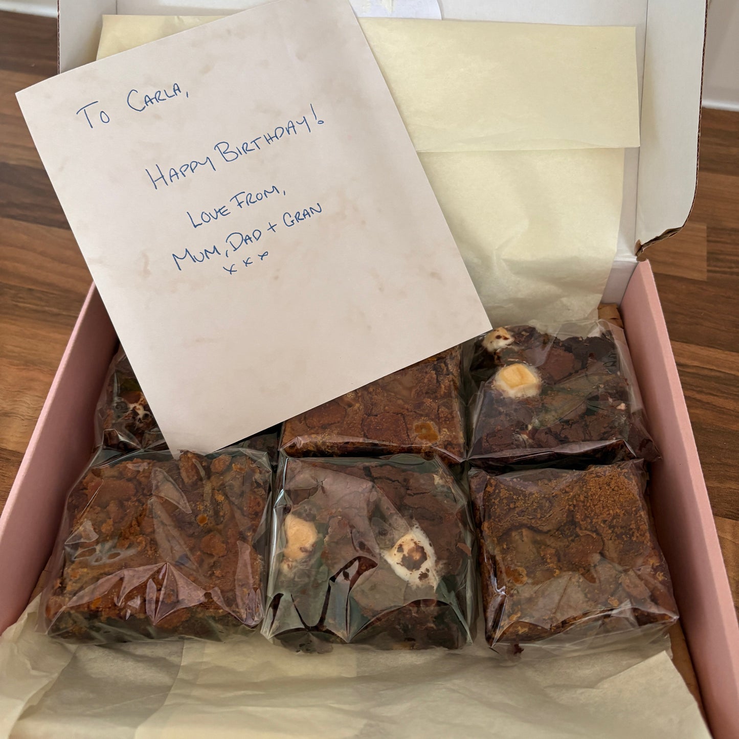 A pink gift box lined with cream tissue paper, containing several individually wrapped brown chocolate treats or cookies. A handwritten note is placed on top of the box.