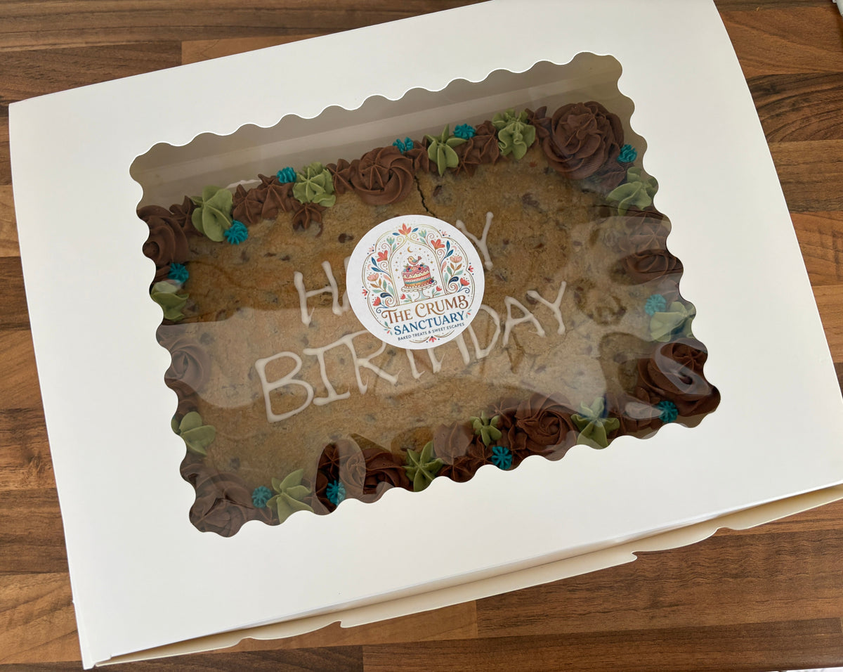 A square white bakery box with a scalloped clear window, showing a chocolate cake inside with piped brown frosting rosettes, green leaf accents, and a round sticker reading 'The Crumb Sanctuary' in the centre. The box is photographed on a wooden surface.