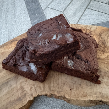 Two rich, glossy chocolate brownies stacked on a rustic live-edge wooden board, set on a gray stone tile.