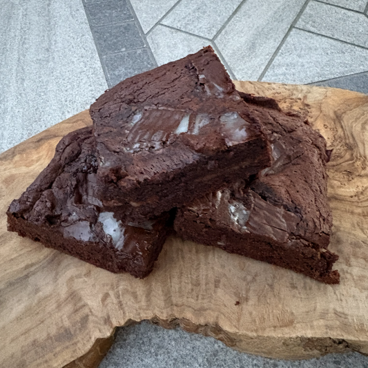 Two rich, glossy chocolate brownies stacked on a rustic live-edge wooden board, set on a gray stone tile.