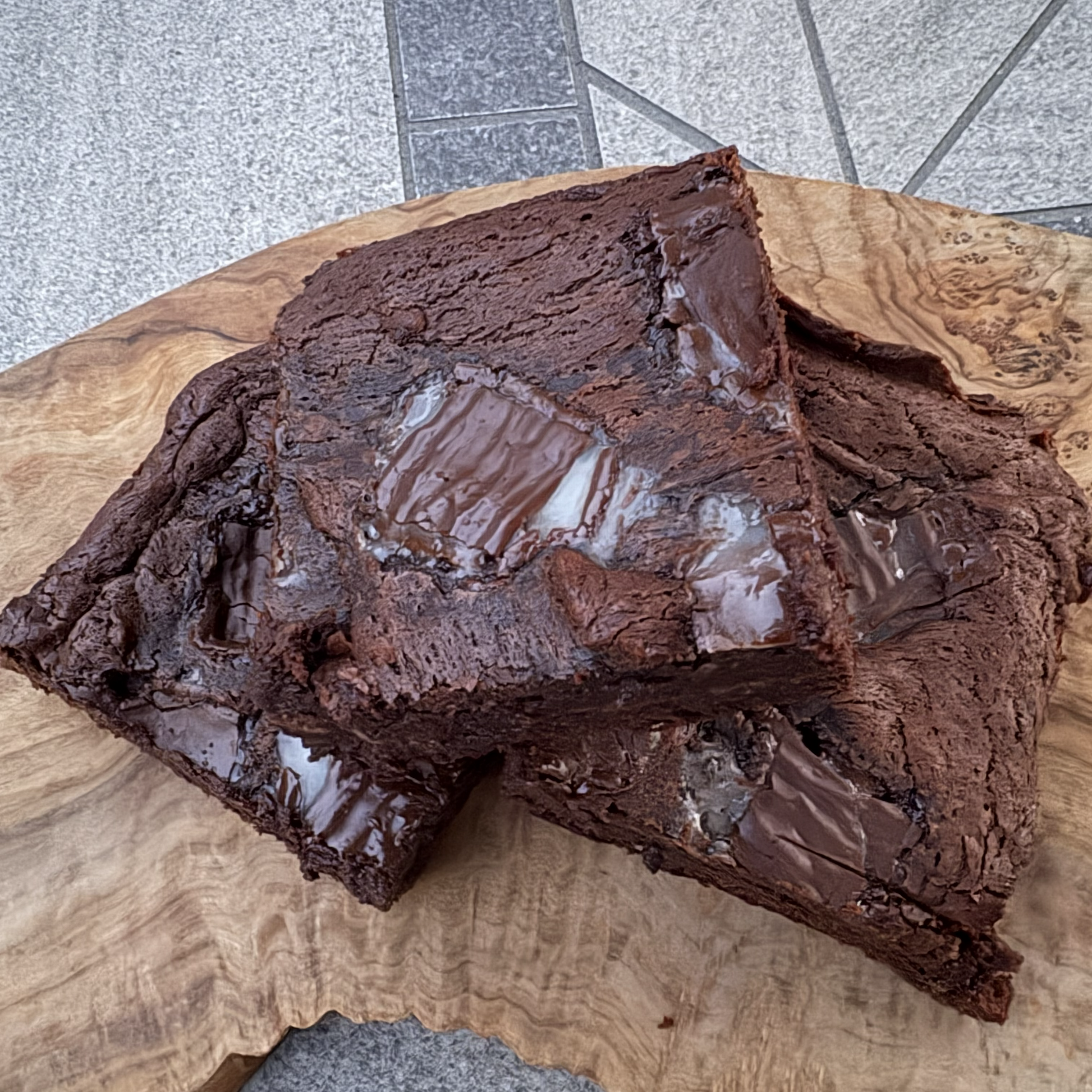 Two rich, glossy chocolate brownies stacked on a rustic live-edge wooden board, set on a gray stone tiled floor.