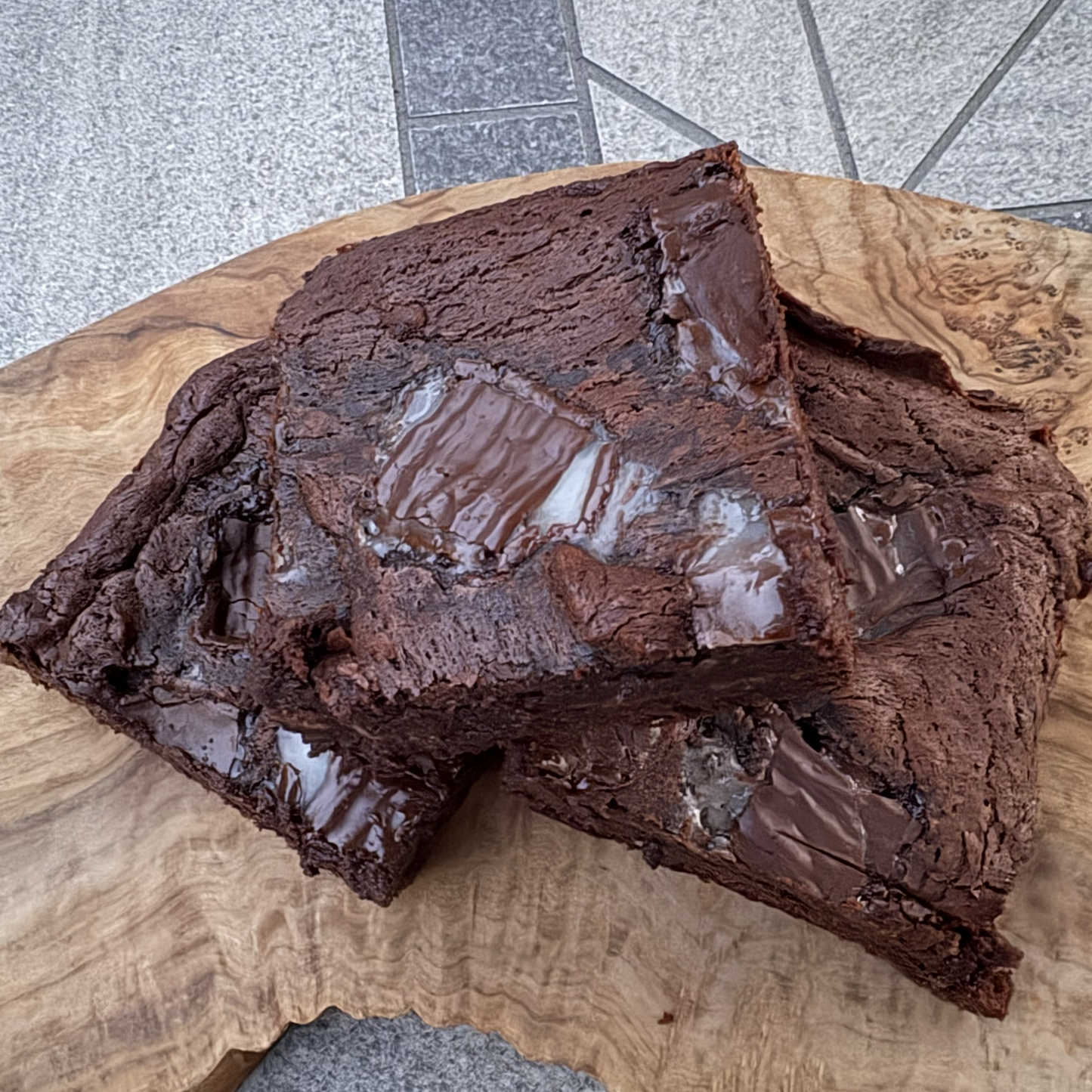 Two rich, glossy chocolate brownies stacked on a rustic live-edge wooden board, set on a gray stone tiled floor.