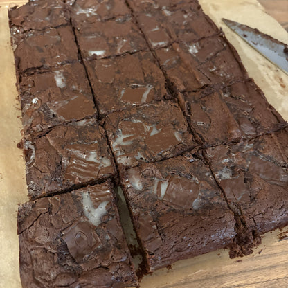A rectangular tray of freshly baked chocolate brownies cut into square portions, with some white icing on top, placed on parchment paper on a wooden surface.