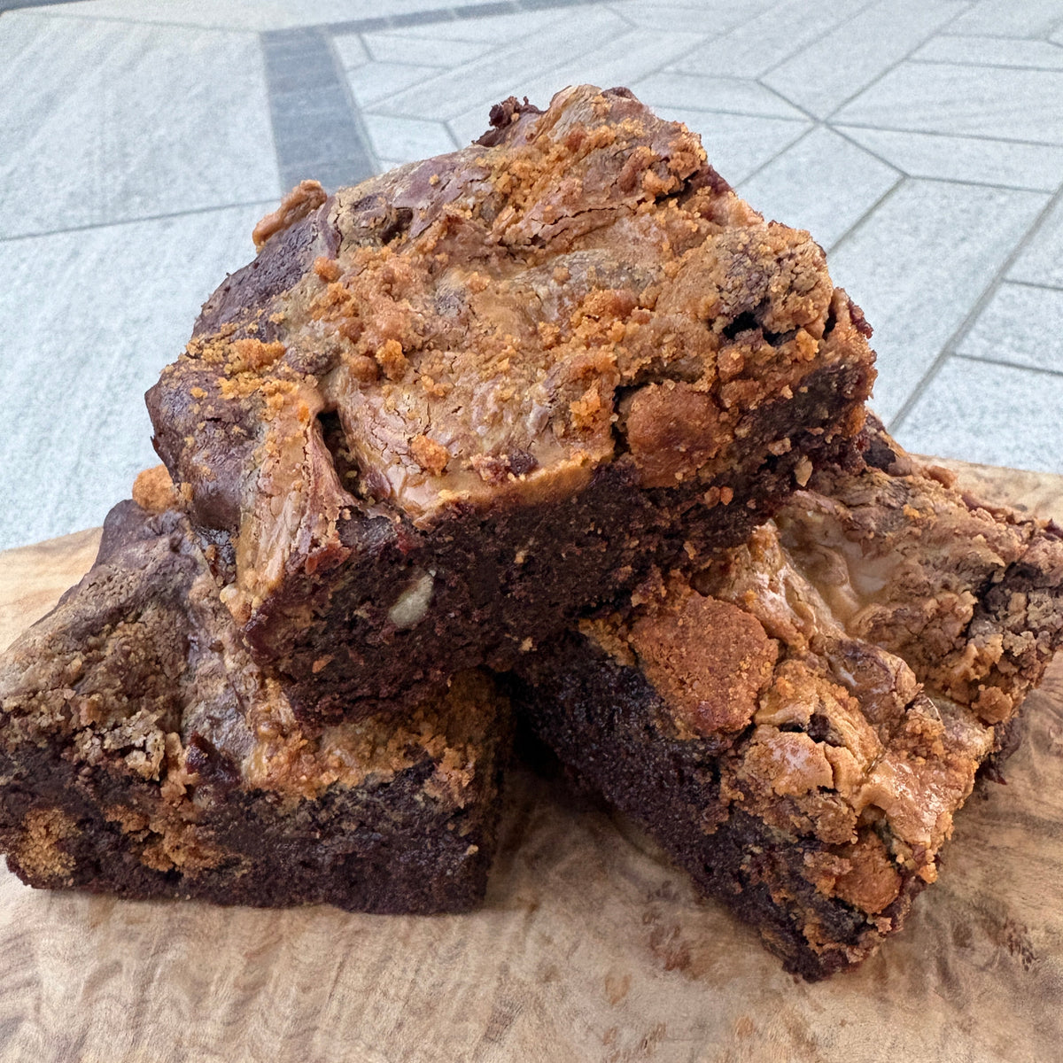 Two freshly baked brownies with a thick crumb and golden-brown topping, presented on a rustic live-edge wooden board over a gray tiled table.
