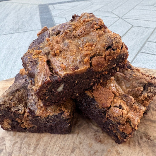 Two freshly baked brownies with a thick crumb and golden-brown topping, presented on a rustic live-edge wooden board over a gray tiled table.