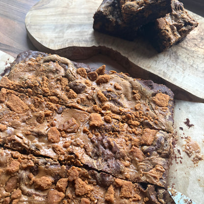A large rectangular brownie or chocolate bar topped with crumbly caramel biscuits and drizzled with Biscoff spread, shown cut into slices on a wooden board with a few stacked pieces in the background.