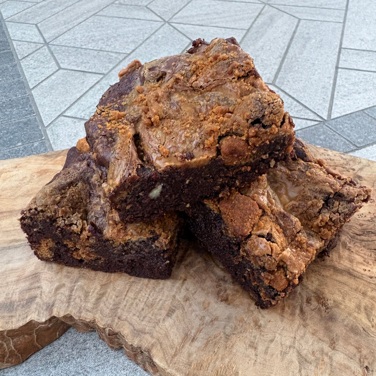 Two rich, dark brownies with golden-brown crumbly tops, stacked on a rustic live-edge wooden board over a gray stone surface.