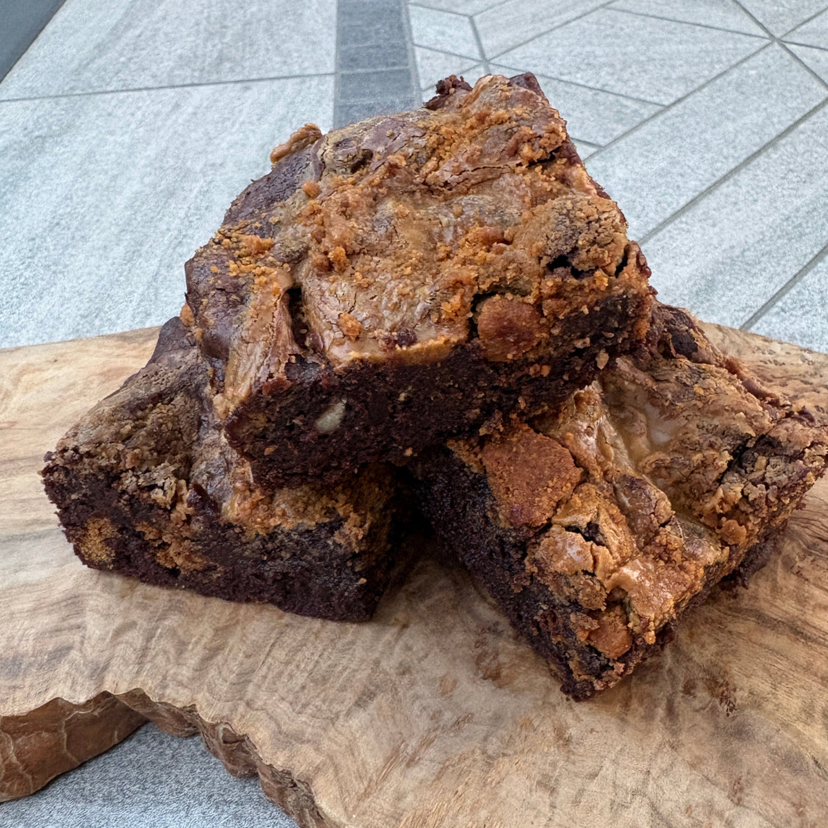 Two rustic brownies with a thick crumb and coarse topping, stacked on a live-edge wooden board over a gray tiled surface.