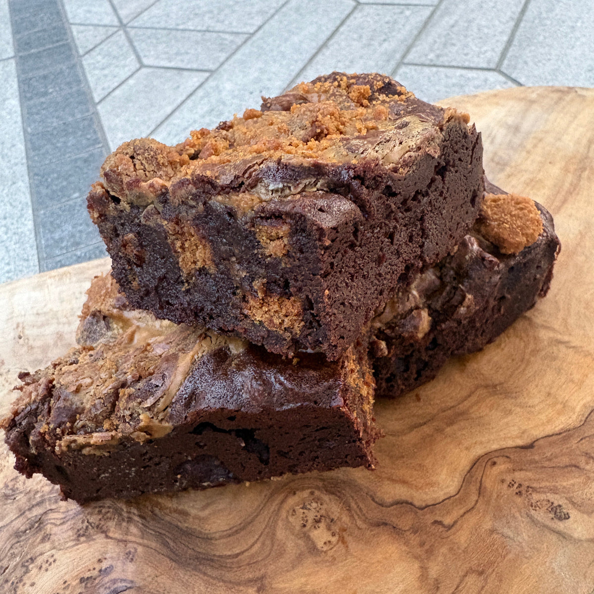 Biscoff brownie with swirls of dark and light brown, sliced and stacked on a large, natural wood cutting board. The background shows a gray tiled table.