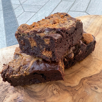 Biscoff brownie with swirls of dark and light brown, sliced and stacked on a large, natural wood cutting board. The background shows a gray tiled table.