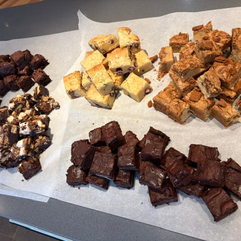 Four different types of sweet treats arranged on parchment paper on a countertop, including Nutella brownies, M+M Blondies, After eight Mint Brownies, and Biscoff blondies.