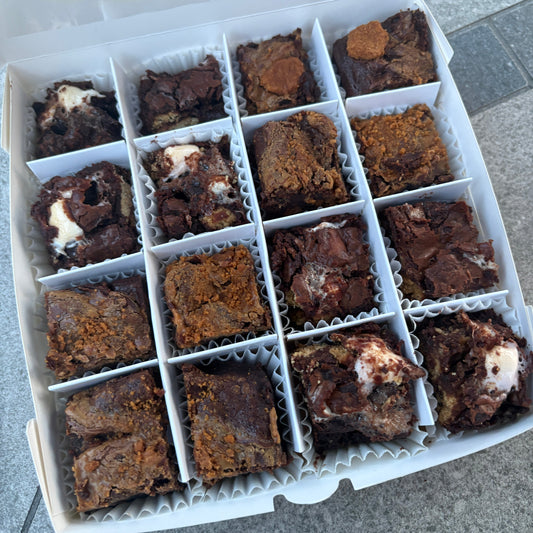 A white gift box with 16 compartments holding assorted chocolate brownies, photographed from above on a grey tiled surface.