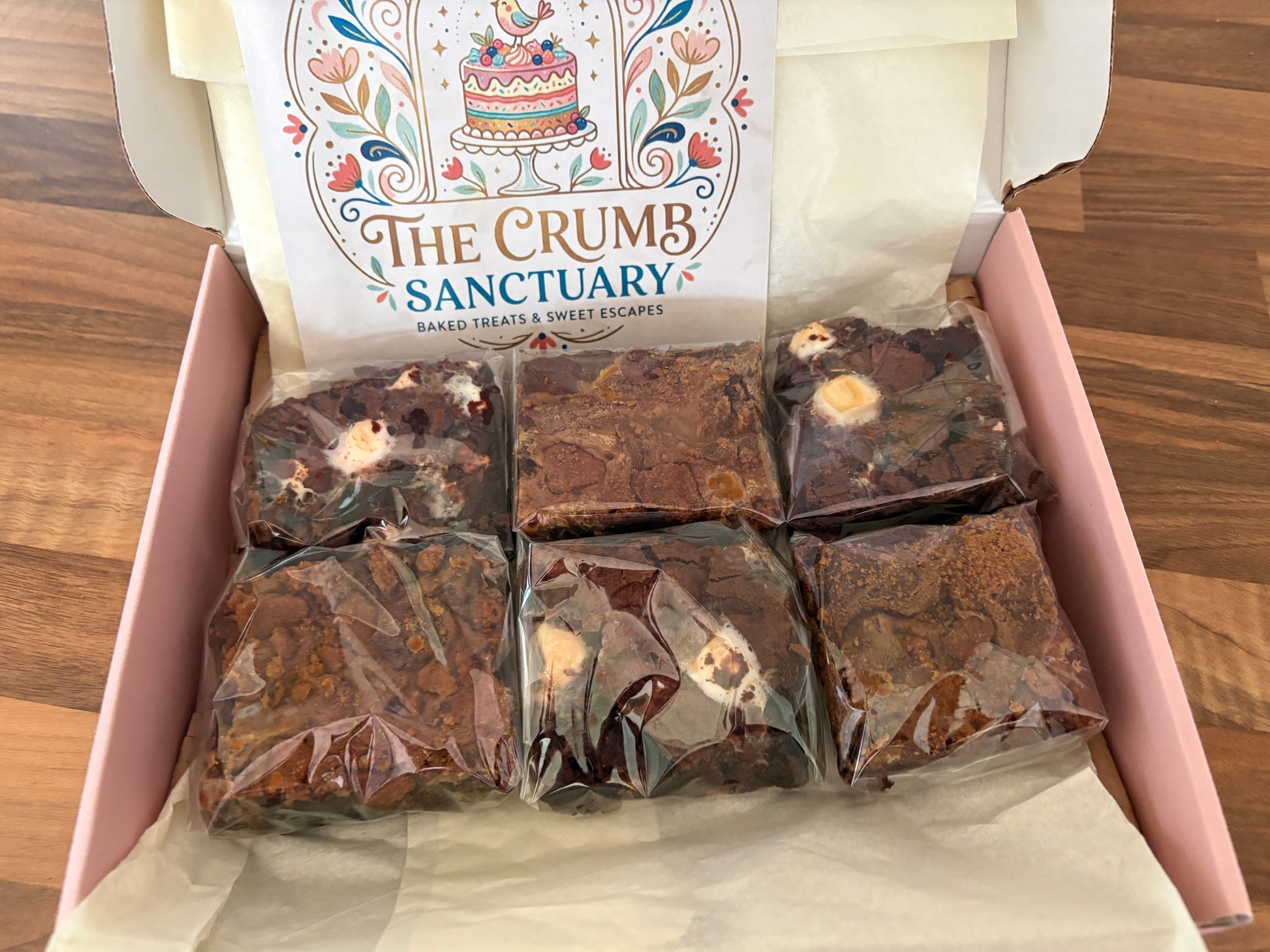 An open pink box of baked goods on a wooden table, showing six individually wrapped brownies or chocolate squares. The inside lid features a printed card reading 'The Crumb Sanctuary'.