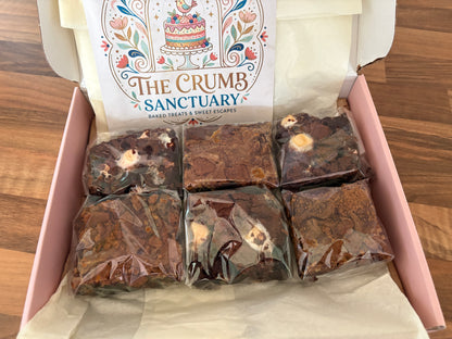 An open pink box of baked goods on a wooden table, showing six individually wrapped brownies or chocolate squares. The inside lid features a printed card reading 'The Crumb Sanctuary'.