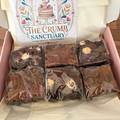 An open pink box of baked goods on a wooden table, showing six individually wrapped brownies or chocolate squares. The inside lid features a printed card reading 'The Crumb Sanctuary'.