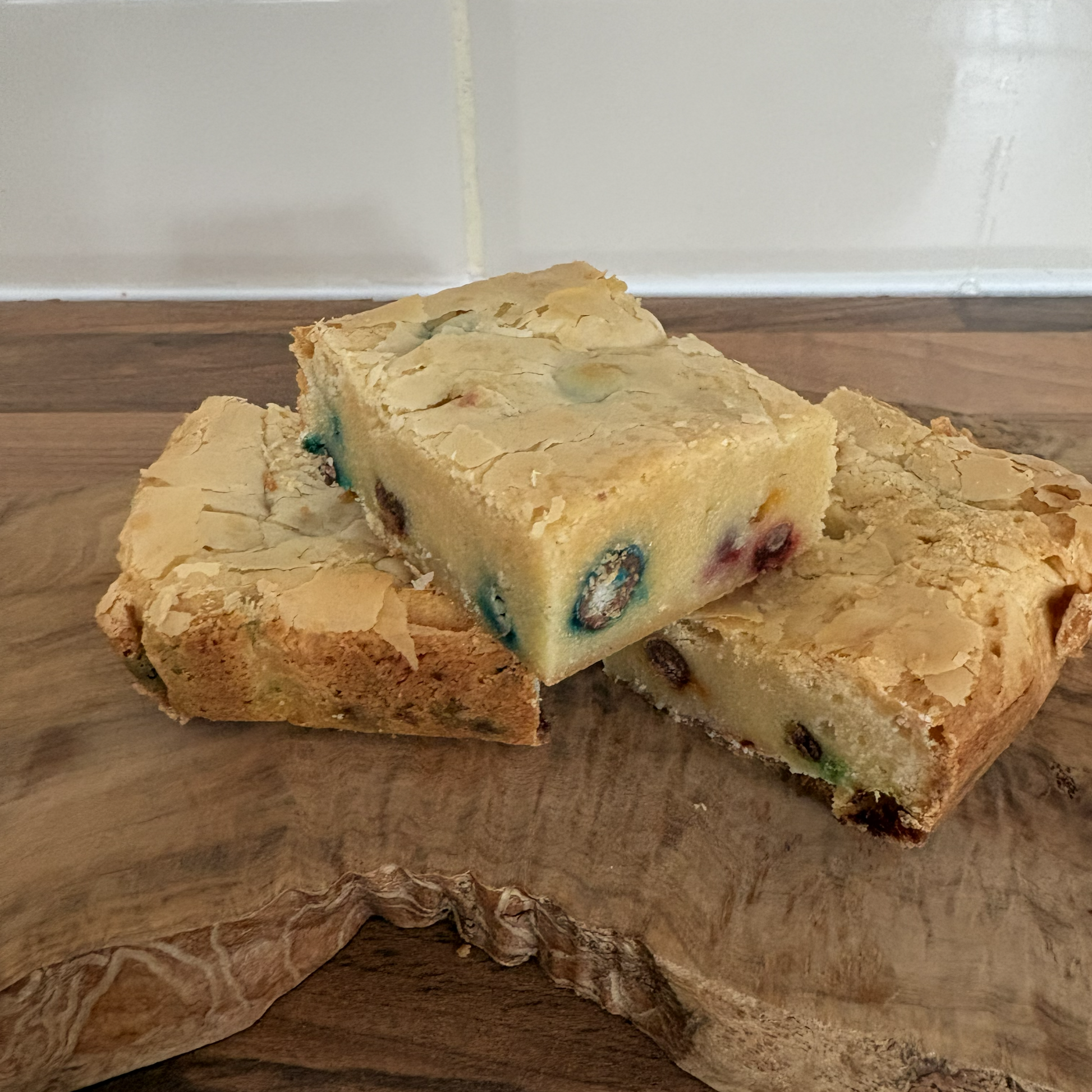 Three pieces of blondie on a wooden cutting board with a tiled wall background