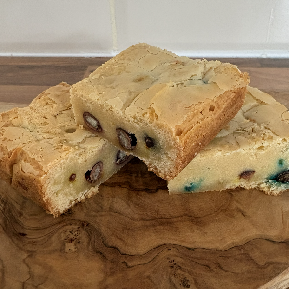 Three Crispy M+M blondies on a wooden cutting board with a tiled backsplash in the background.