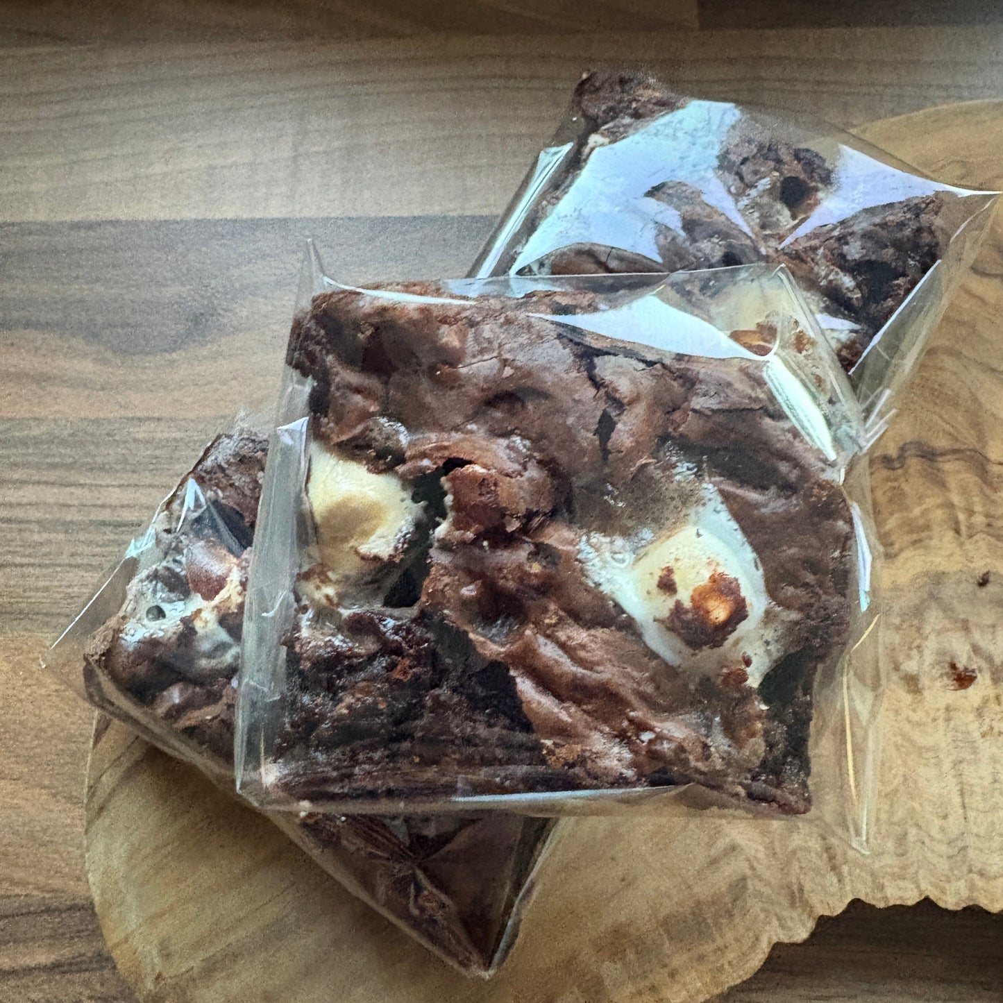 Three square brownies with white chocolate swirls and toasted marshmallow pieces, individually wrapped in clear plastic and stacked on a wooden board.