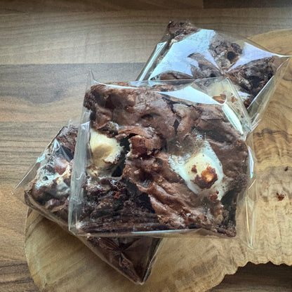 Three square brownies with white chocolate swirls and toasted marshmallow pieces, individually wrapped in clear plastic and stacked on a wooden board.
