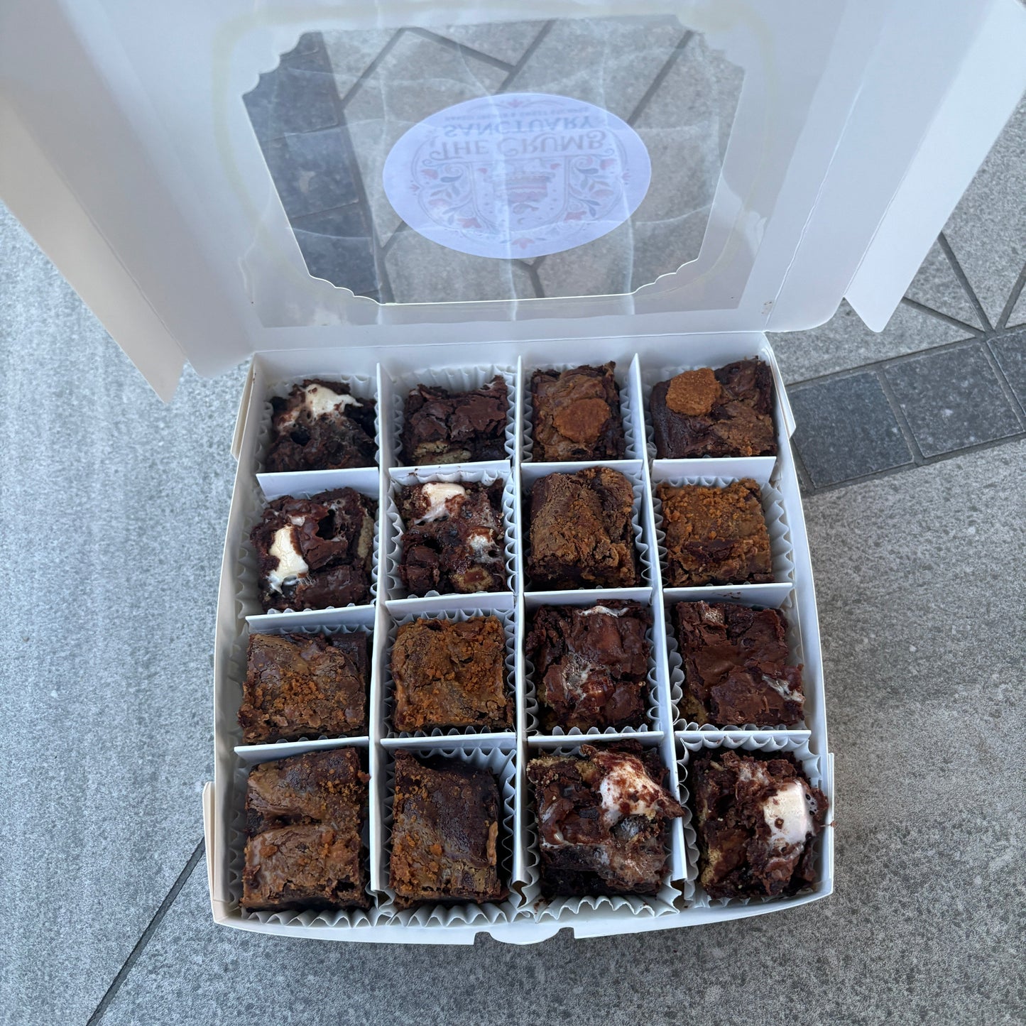 An open white cardboard box containing 16 assorted chocolate brownies, photographed from above on a grey surface.