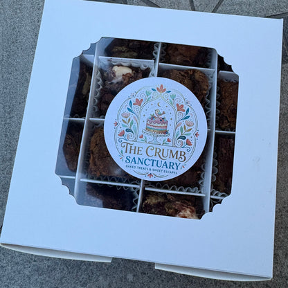 A white cardboard gift box with a window displaying assorted brown baked goods, featuring a round printed label that reads 'The Crumb Sanctuary'.