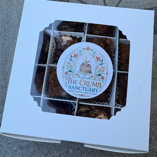 A white cardboard gift box with a window displaying assorted brown baked goods, featuring a round printed label that reads 'The Crumb Sanctuary'.
