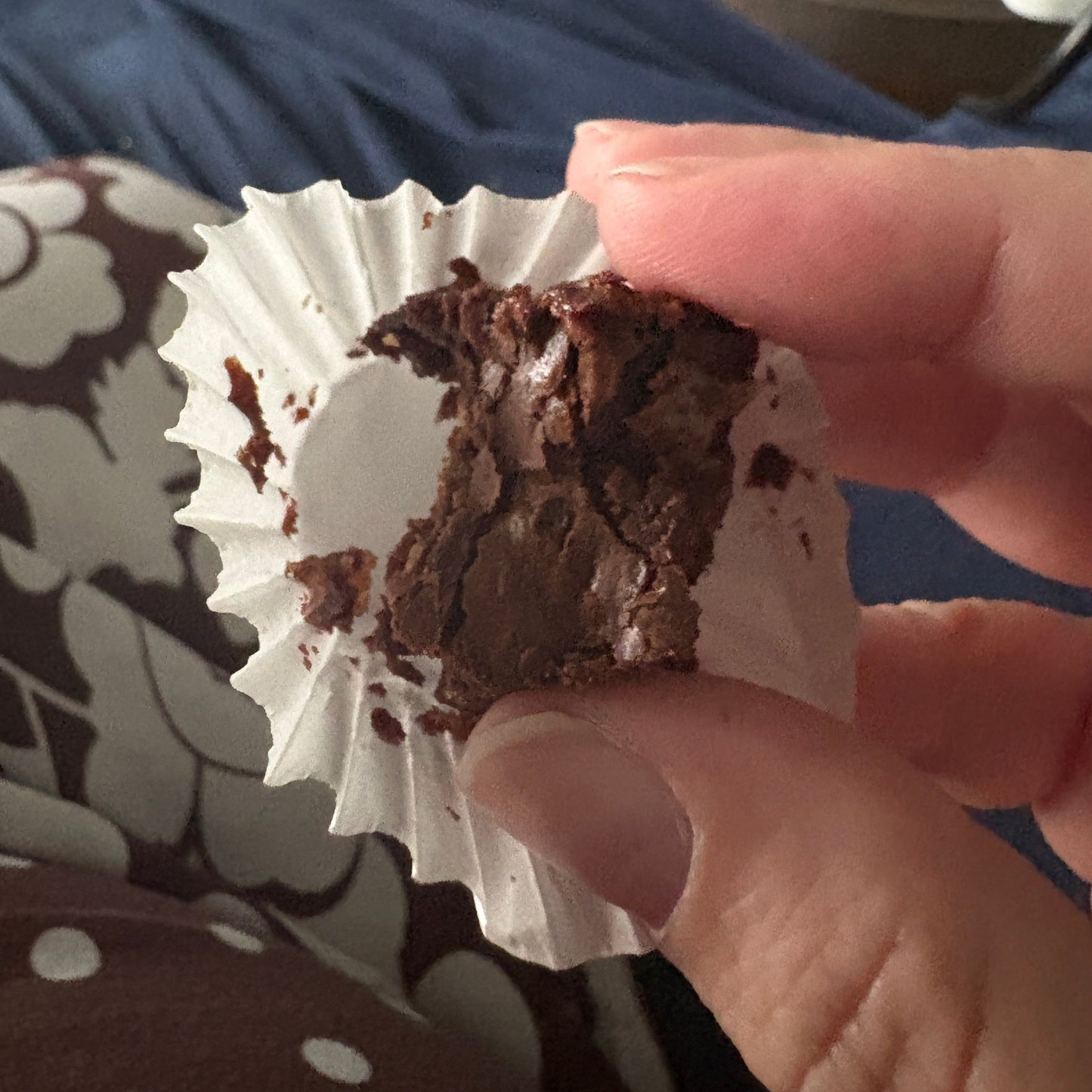 A close-up of a hand holding a partially eaten chocolate brownie in a white paper liner, with a patterned blanket and blue clothing in the background.