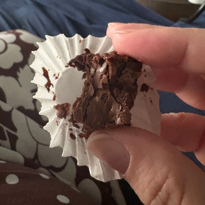 A close-up of a hand holding a partially eaten chocolate brownie in a white paper liner, with a patterned blanket and blue clothing in the background.