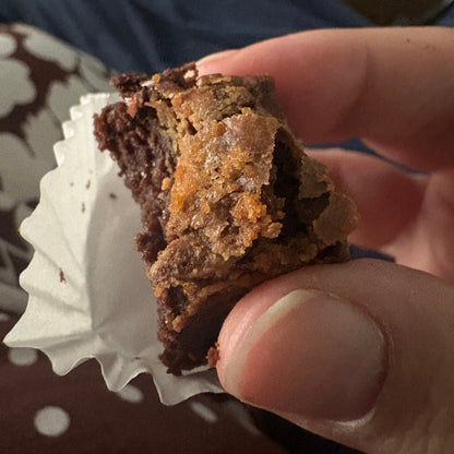A close-up of a hand holding a bite-sized brownie with a white paper liner, set against a background of dark blue clothing and a brown patterned surface.