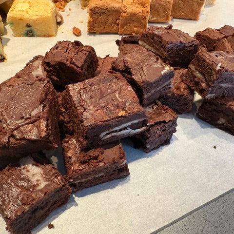 A pile of freshly cut chocolate brownies with various toppings, including white chocolate drizzle, nuts, and cream, arranged on a light surface with other baked goods blurred in the background.
