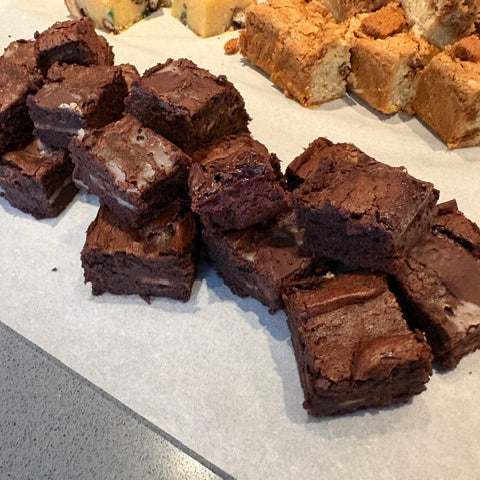 A close-up shot of freshly baked chocolate brownies stacked on white parchment paper, with other assorted baked goods blurred in the background.