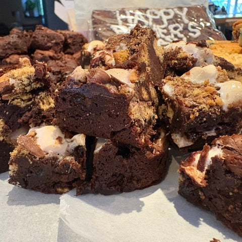 A close-up pile of rich chocolate brownies with white chocolate drizzle, nuts, and cookie pieces, arranged on parchment paper with other baked goods blurred in the background.
