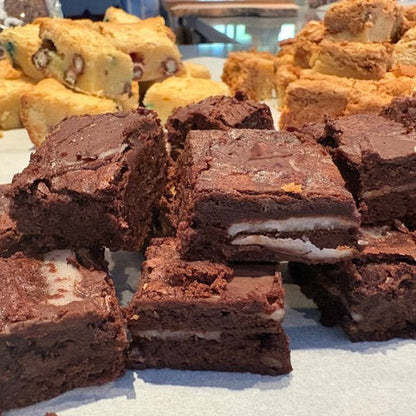 A close-up of assorted chocolate brownies stacked on a white surface, with other colorful baked goods blurred in the background.