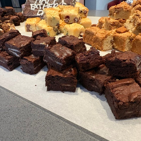 An assortment of baked goods displayed on a counter, featuring chocolate brownies in the foreground, golden-brown sugar squares on the right, and other assorted sweets in the background.