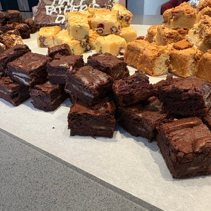 An assortment of baked goods displayed on a counter, featuring chocolate brownies in the foreground, golden-brown sugar squares on the right, and other assorted sweets in the background.
