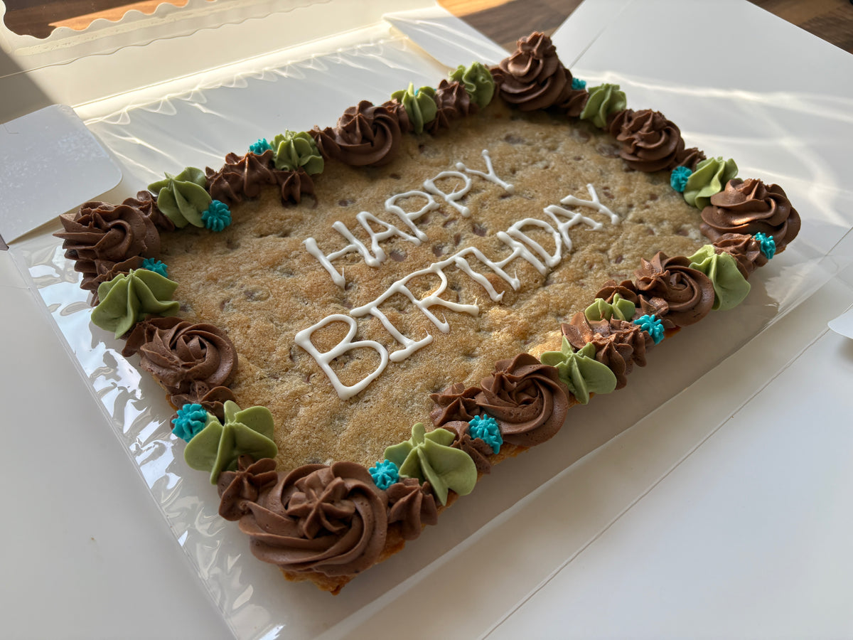 A rectangular birthday cookie cake decorated with white 'HAPPY BIRTHDAY' icing and a border of brown and green frosting rosettes, sitting in a clear plastic container.