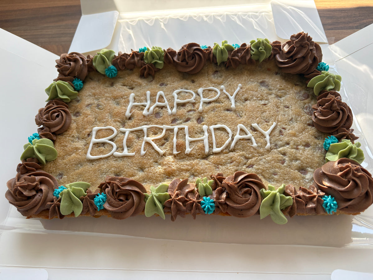A rectangular birthday cookie cake in a white box, decorated with brown frosting rosettes, green and blue accents, and white icing writing that says 'HAPPY BIRTHDAY'.