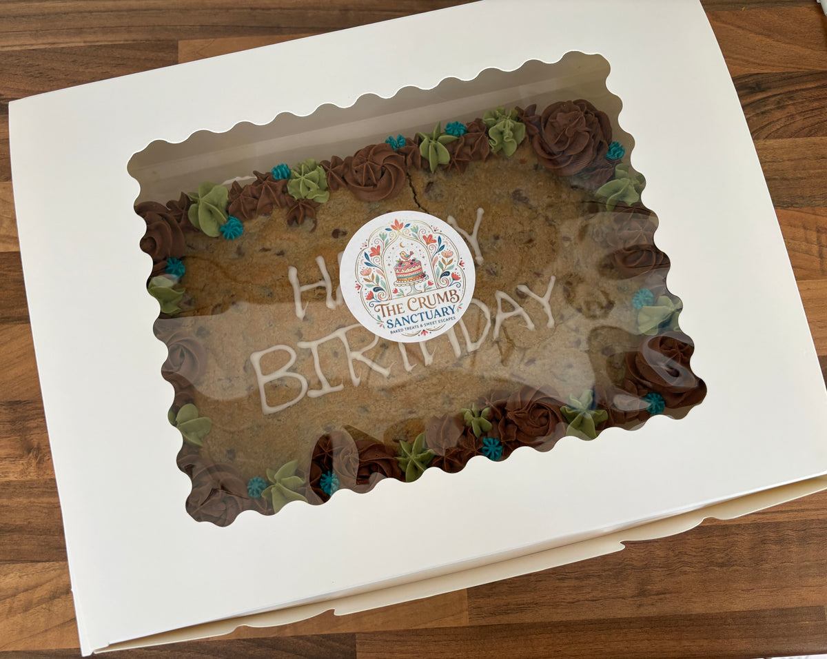 A square white bakery box with a scalloped clear window, showing a chocolate cake inside with piped brown frosting rosettes, green leaf accents, and a round sticker reading 'The Crumb Sanctuary' in the centre. The box is photographed on a wooden surface.