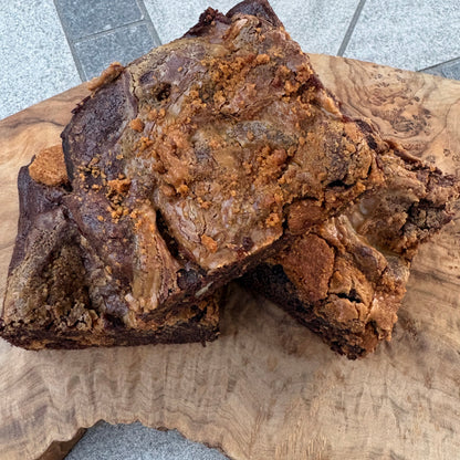 Three large biscoff chocolate brownie slices displayed on a live-edge wooden board over a gray table.
