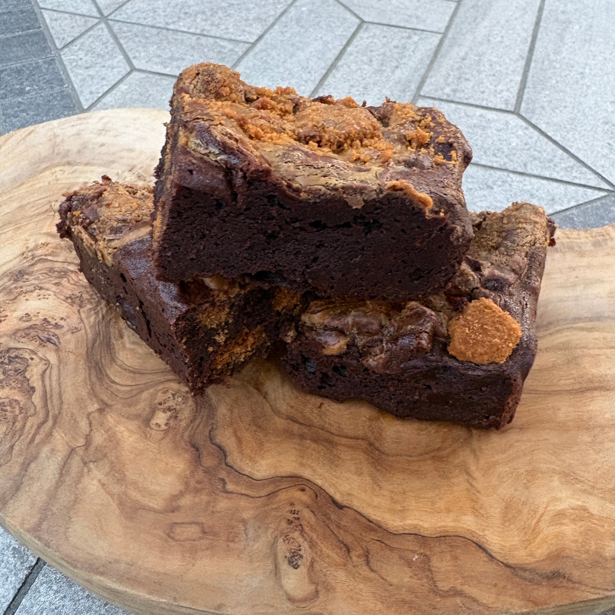 A close-up of a dark chocolate brownie with a crumbly golden-brown topping, resting on a round natural wood slab with a gray tiled floor visible in the background.