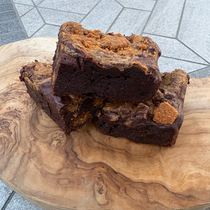 A close-up of a dark chocolate brownie with a crumbly golden-brown topping, resting on a round natural wood slab with a gray tiled floor visible in the background.