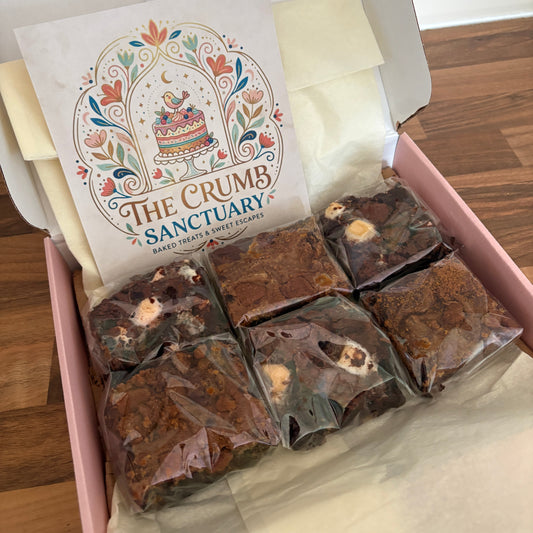 A gift box of artisanal chocolate squares from The Crumb's Chocolate Company, shown in a pink box with tissue paper and a branded card on a wooden floor background.