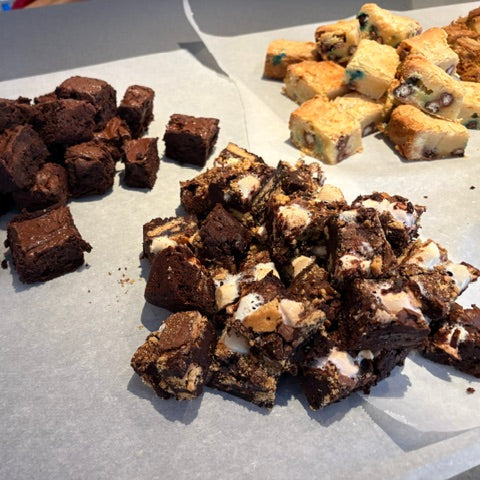 Three different types of baked dessert bars arranged on parchment paper: chocolate Nutella brownies, rock road brownies, and M+M Blondies