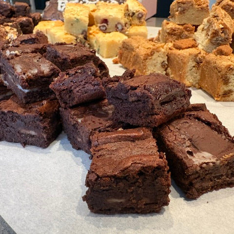 A close-up display of freshly baked brownies in the foreground, with various colorful dessert bars and pieces arranged in the background on a white surface.