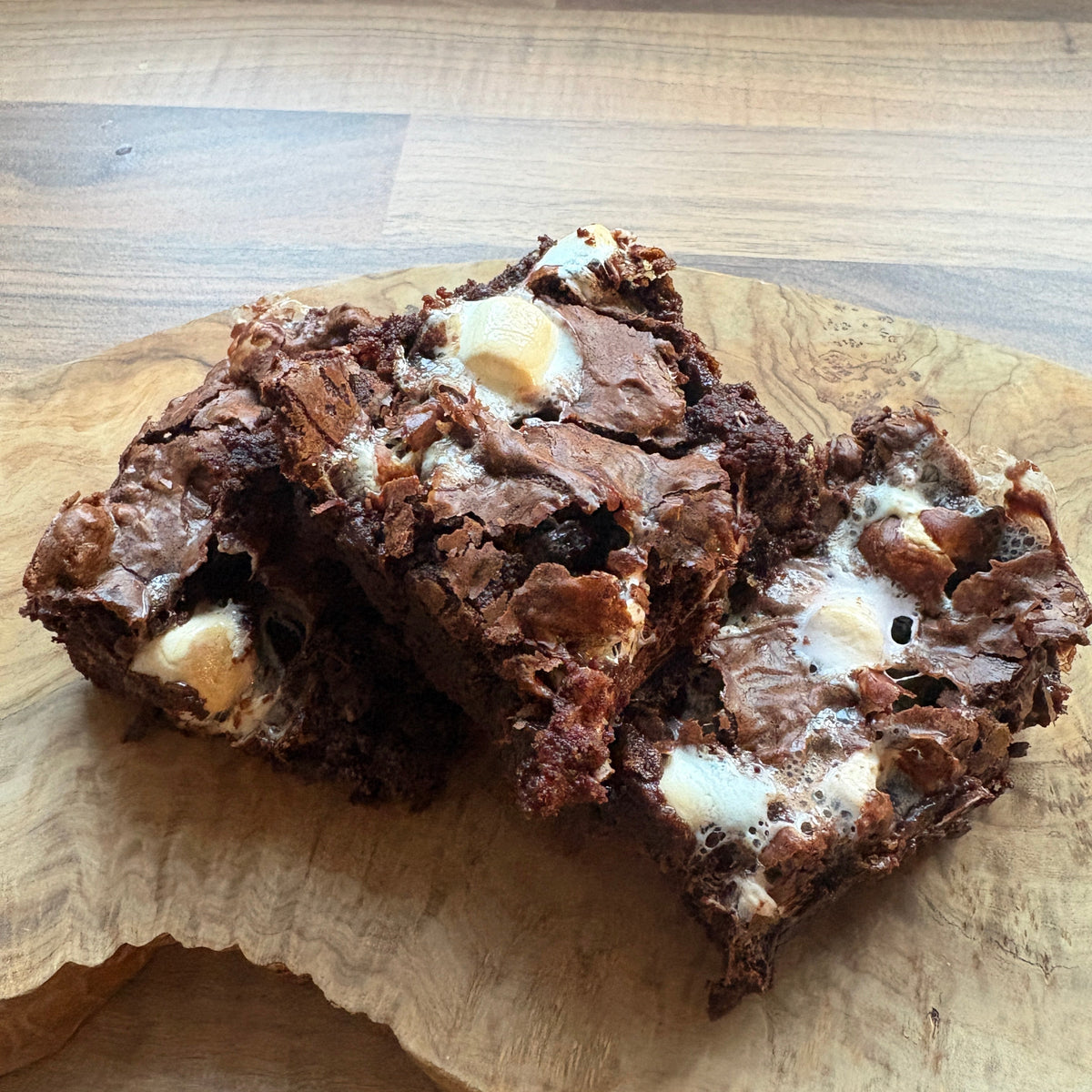 Three chocolate brownies with white chocolate chips stacked on a wooden serving board over a wood floor.