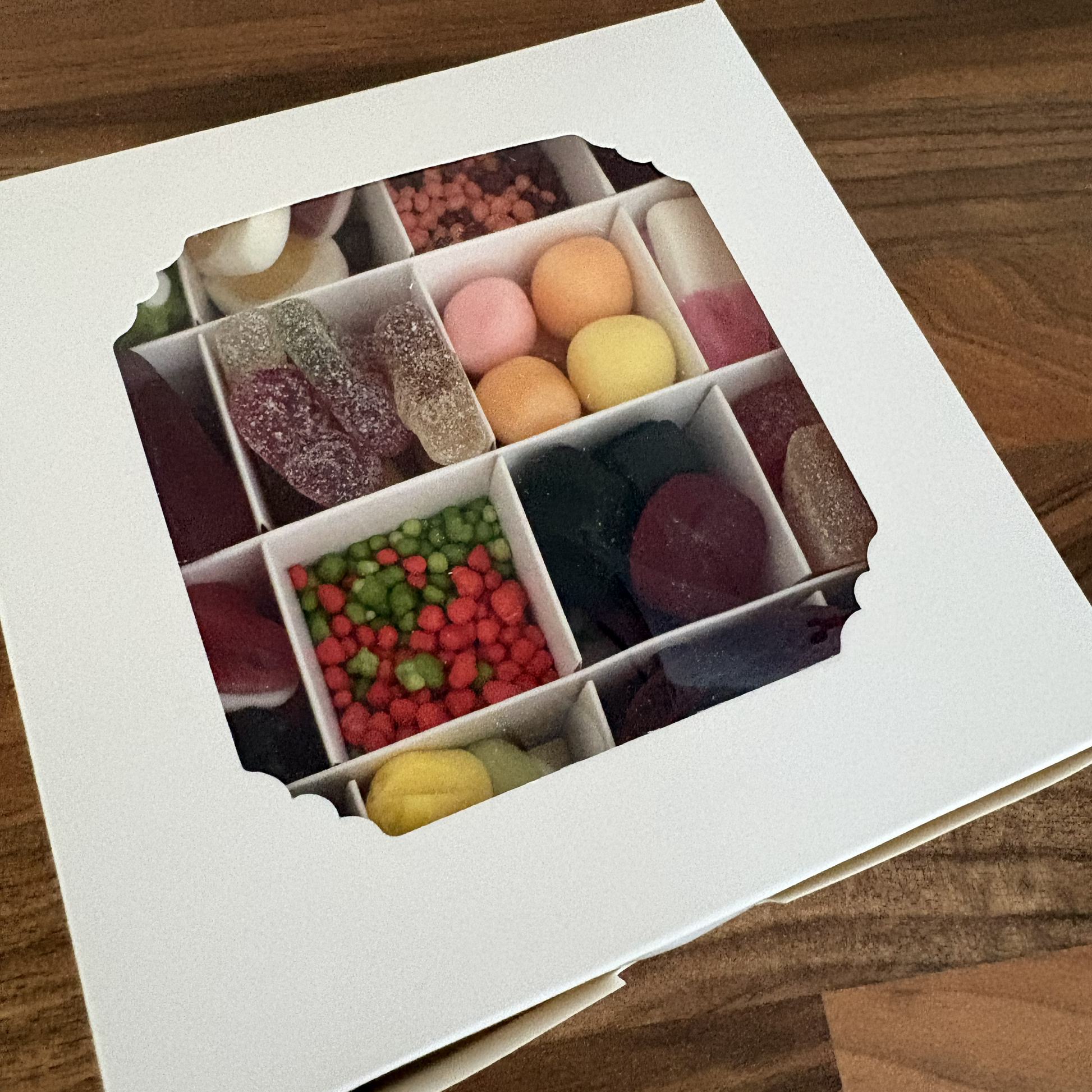 A white square gift box with a window showing a 3x3 grid of assorted colorful candies, gummy sweets, and hard candies, placed on a wooden tabletop.