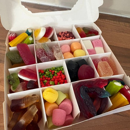 An open white cardboard box filled with assorted colorful candies and gummy sweets, photographed from above on a wooden surface.