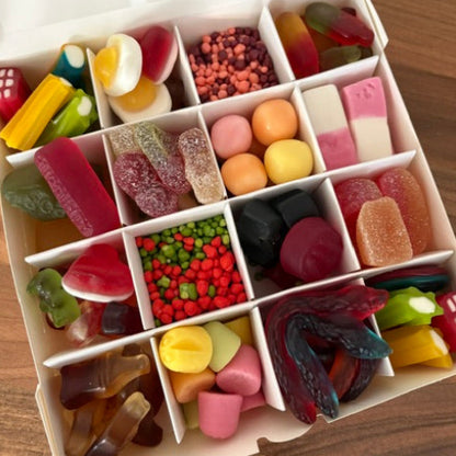An open white candy box filled with assorted gummy candies, hard candies, and sugar-coated sweets arranged in compartments, photographed from above on a wooden surface.