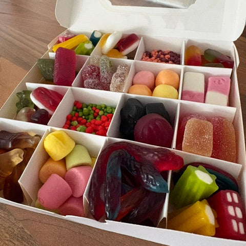 A white cardboard box filled with assorted colorful gummy candies and sweets, photographed from above on a wooden surface.